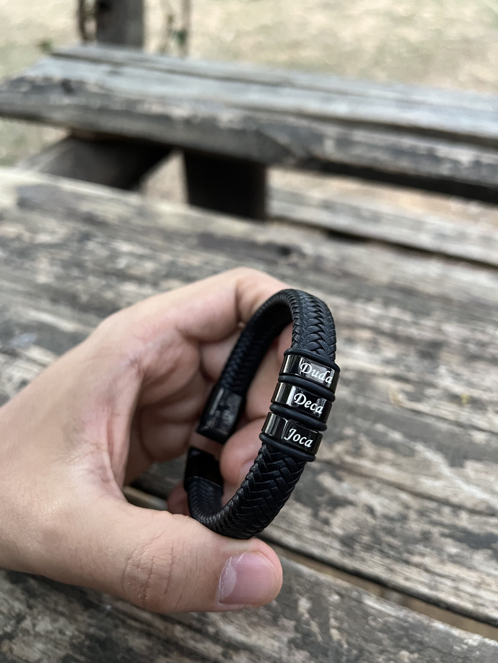 Mão segurando pulseira masculina de couro trançado preto com contas gravadas Duda, Deca e Joca